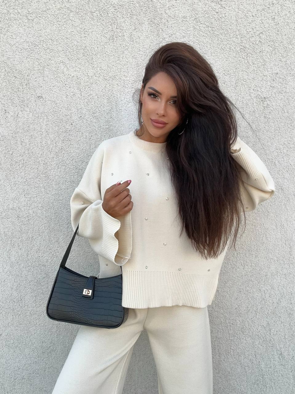 Woman wearing a white sweater and pants with a black handbag against a light gray wall.