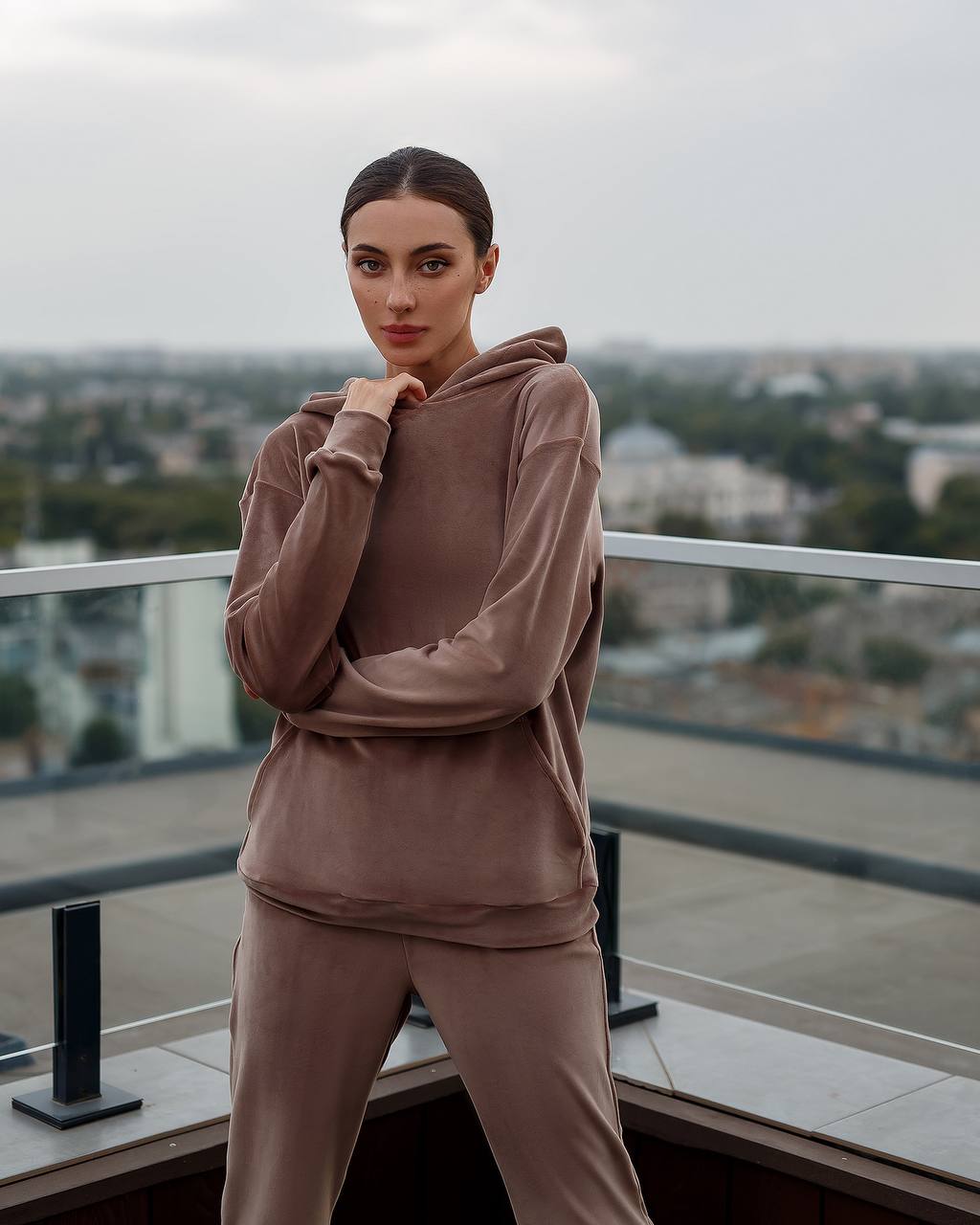 Woman in a brown tracksuit standing on a balcony with a cityscape background