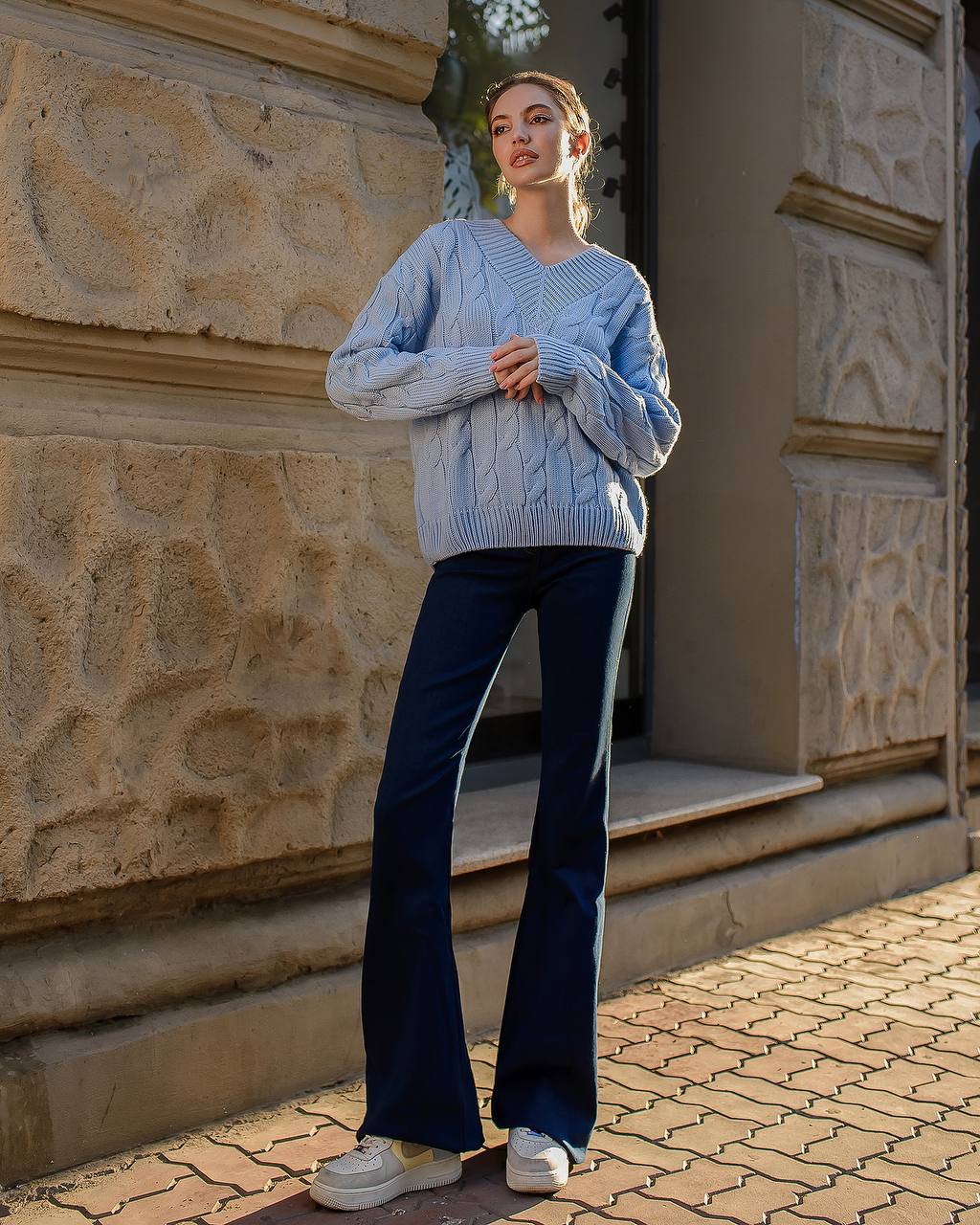 Person wearing a light blue cable knit sweater and dark blue flared jeans standing in front of a stone building.