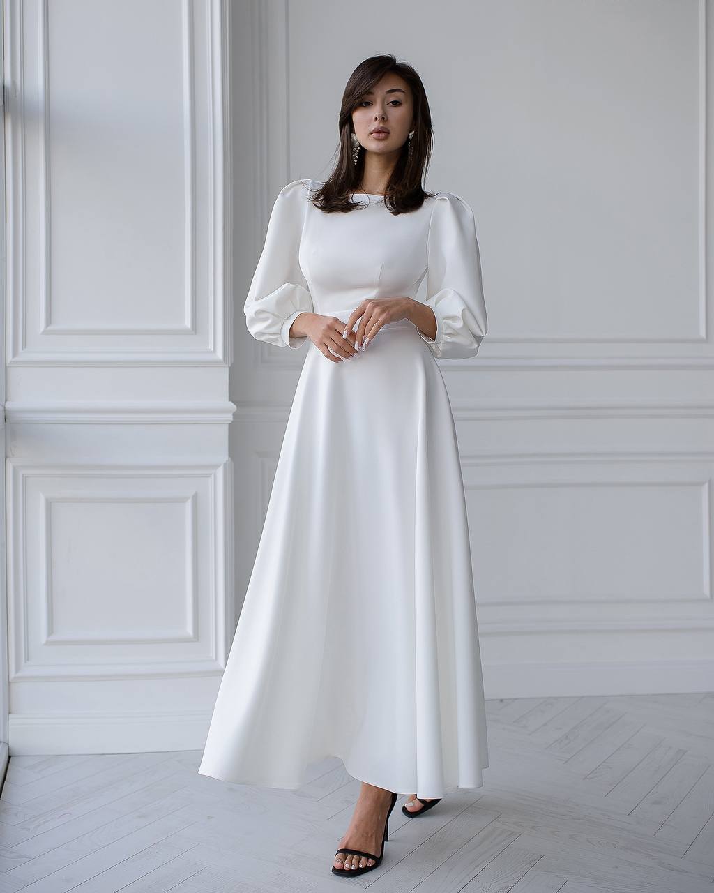 Woman wearing a white dress standing against a white wall.