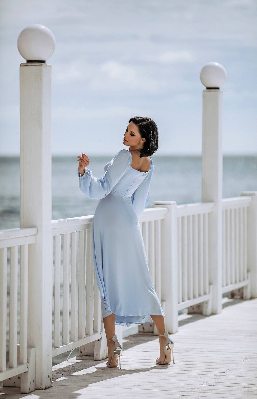 Woman in a light blue dress standing on a wooden boardwalk by the sea.