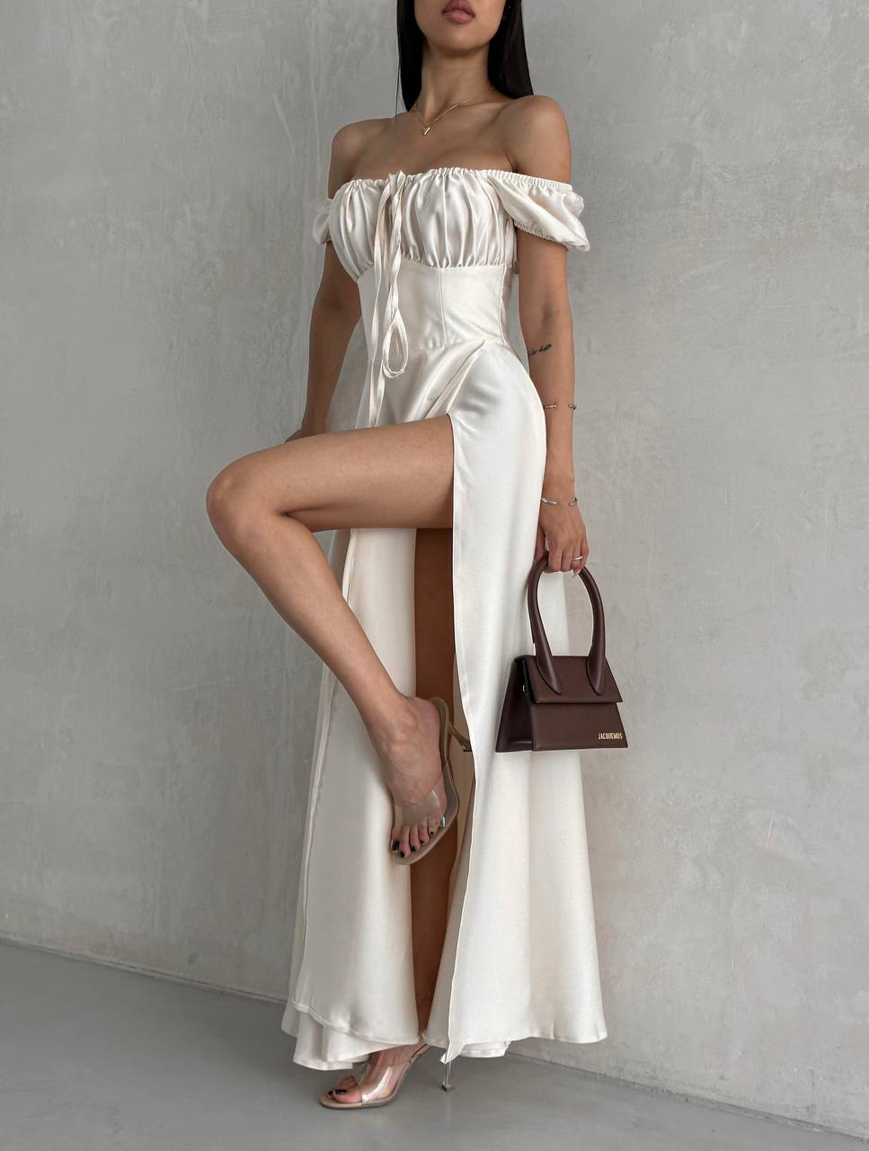 Woman wearing a white off-shoulder dress with a brown handbag against a gray background