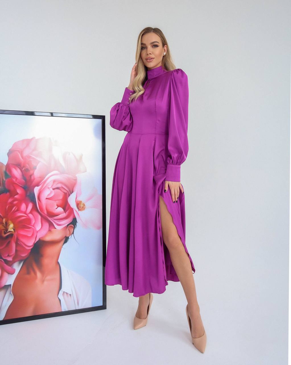 Woman in a purple dress standing next to a framed floral artwork on a white wall.