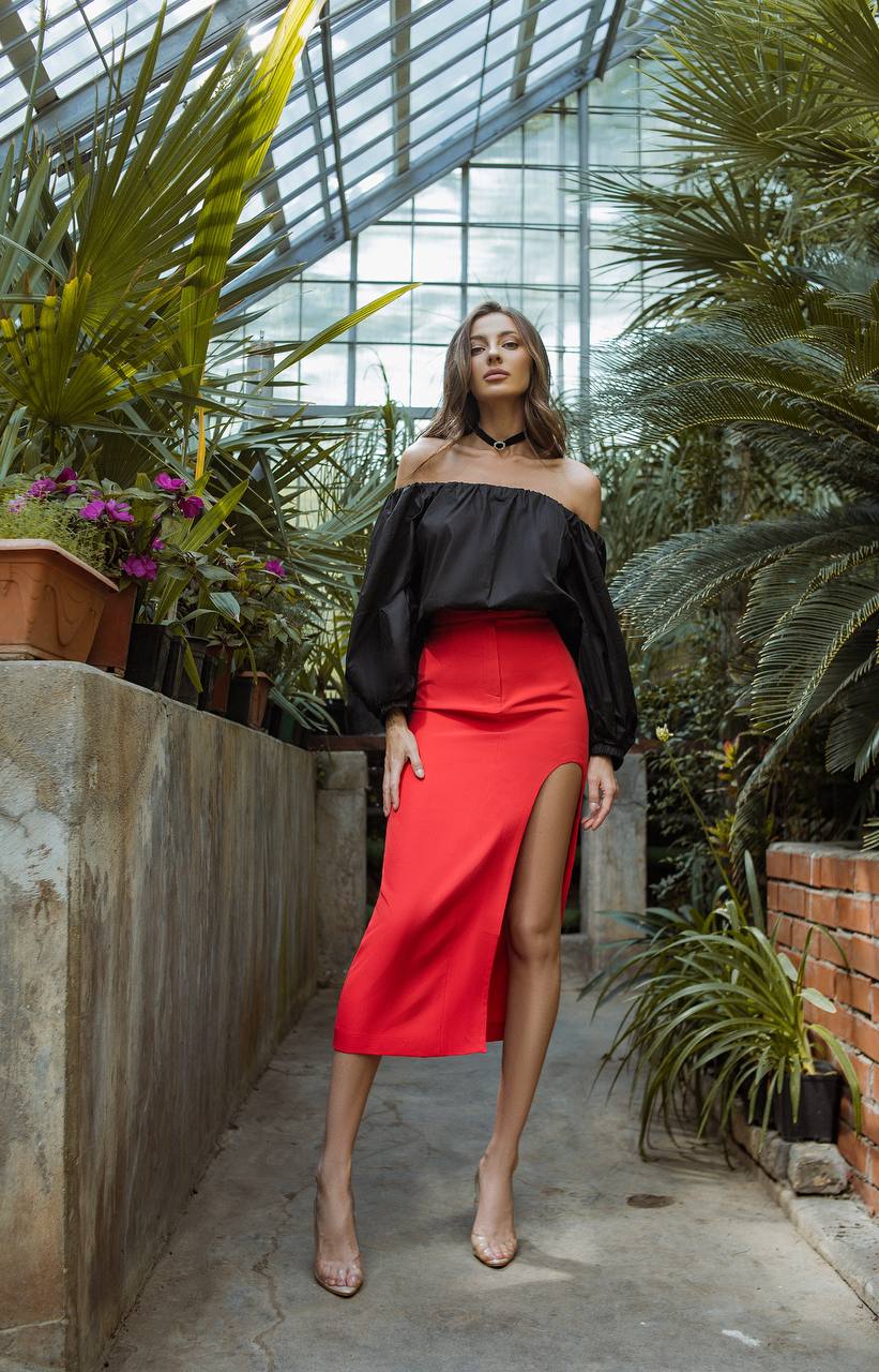 Woman in a black top and red skirt standing in a greenhouse with plants around.