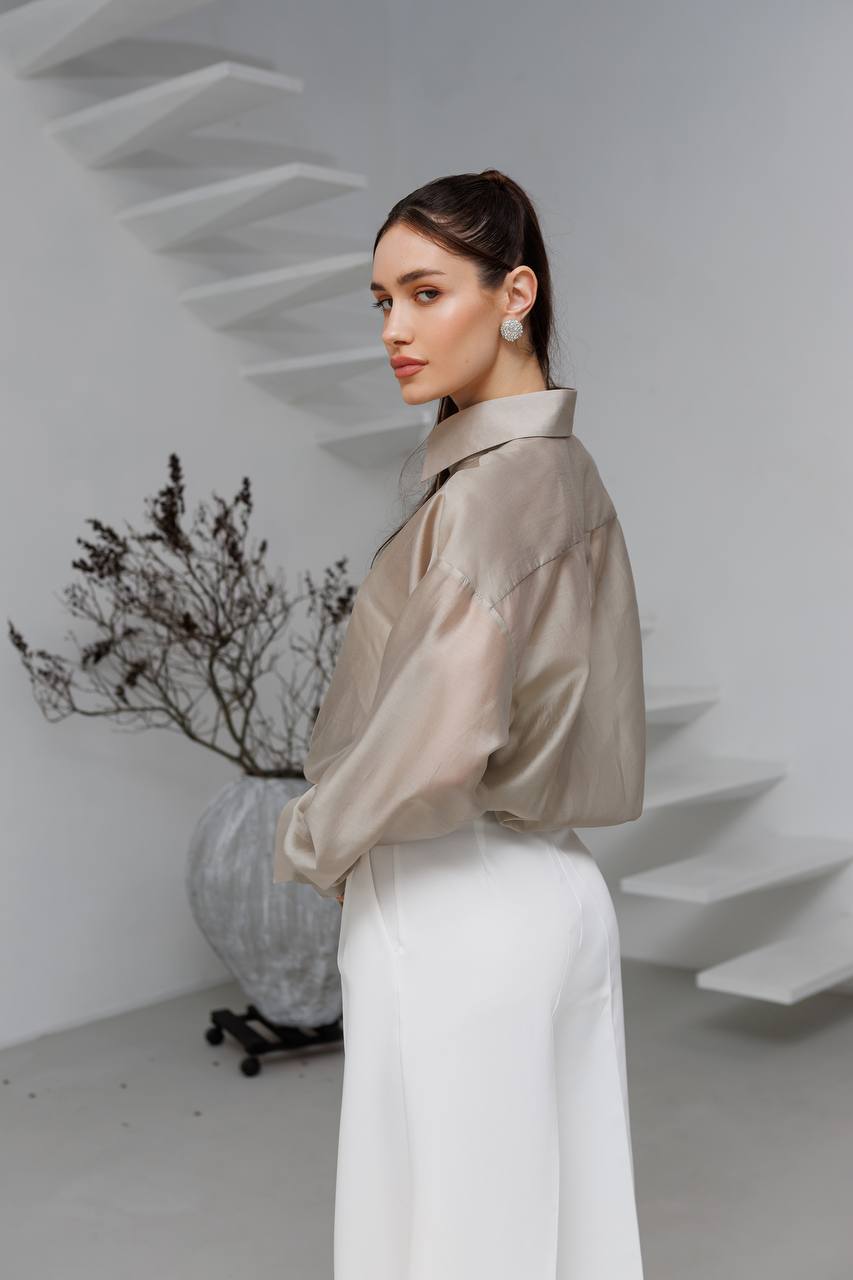 Woman in a beige blouse and white pants standing in a minimalistic room with a plant.