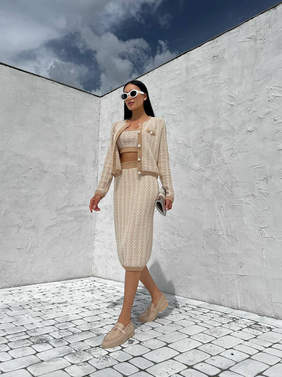 Woman in a beige outfit standing against a white wall with a blue sky.