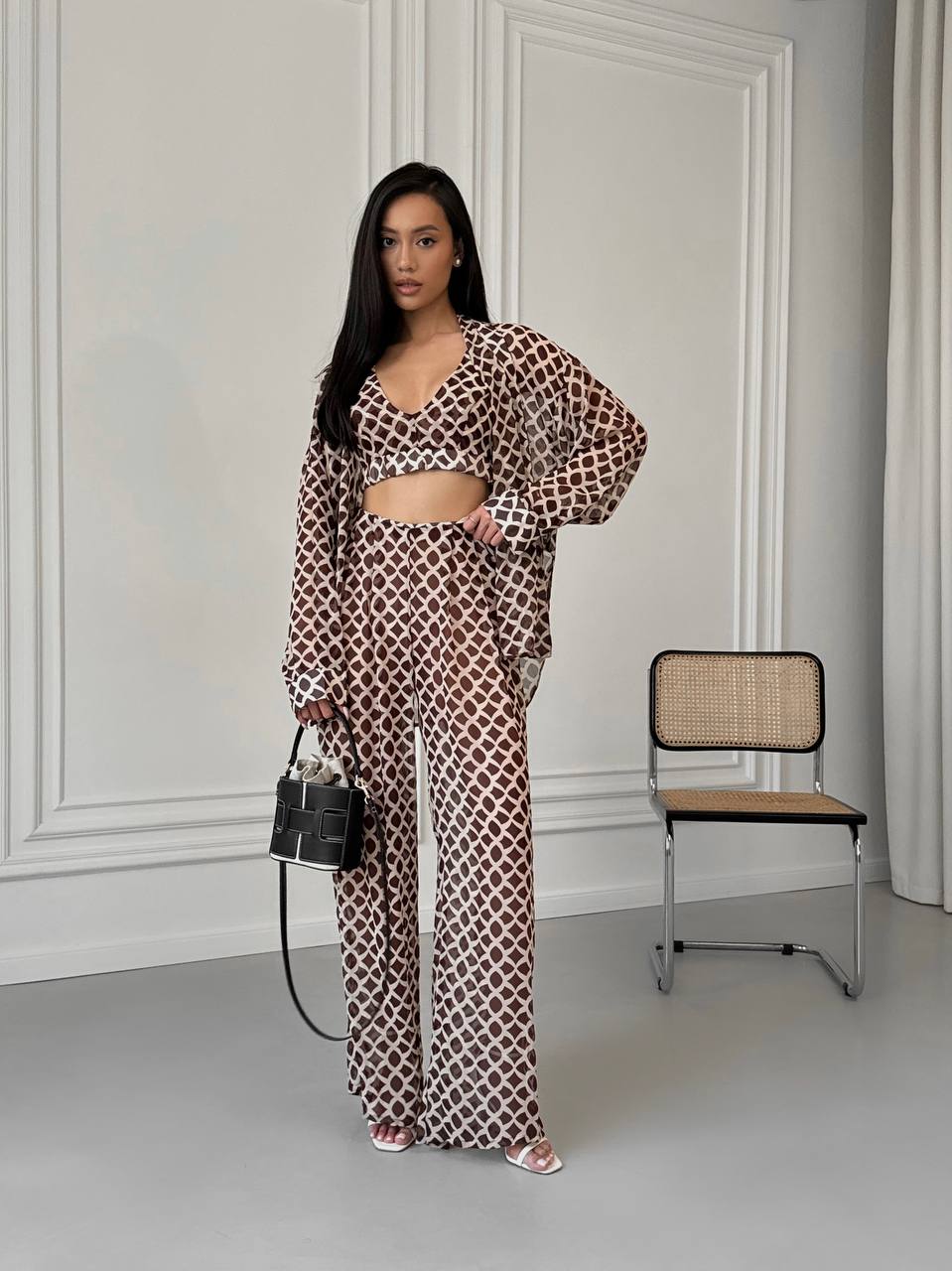 Woman in a patterned outfit standing in a room with a chair and handbag.