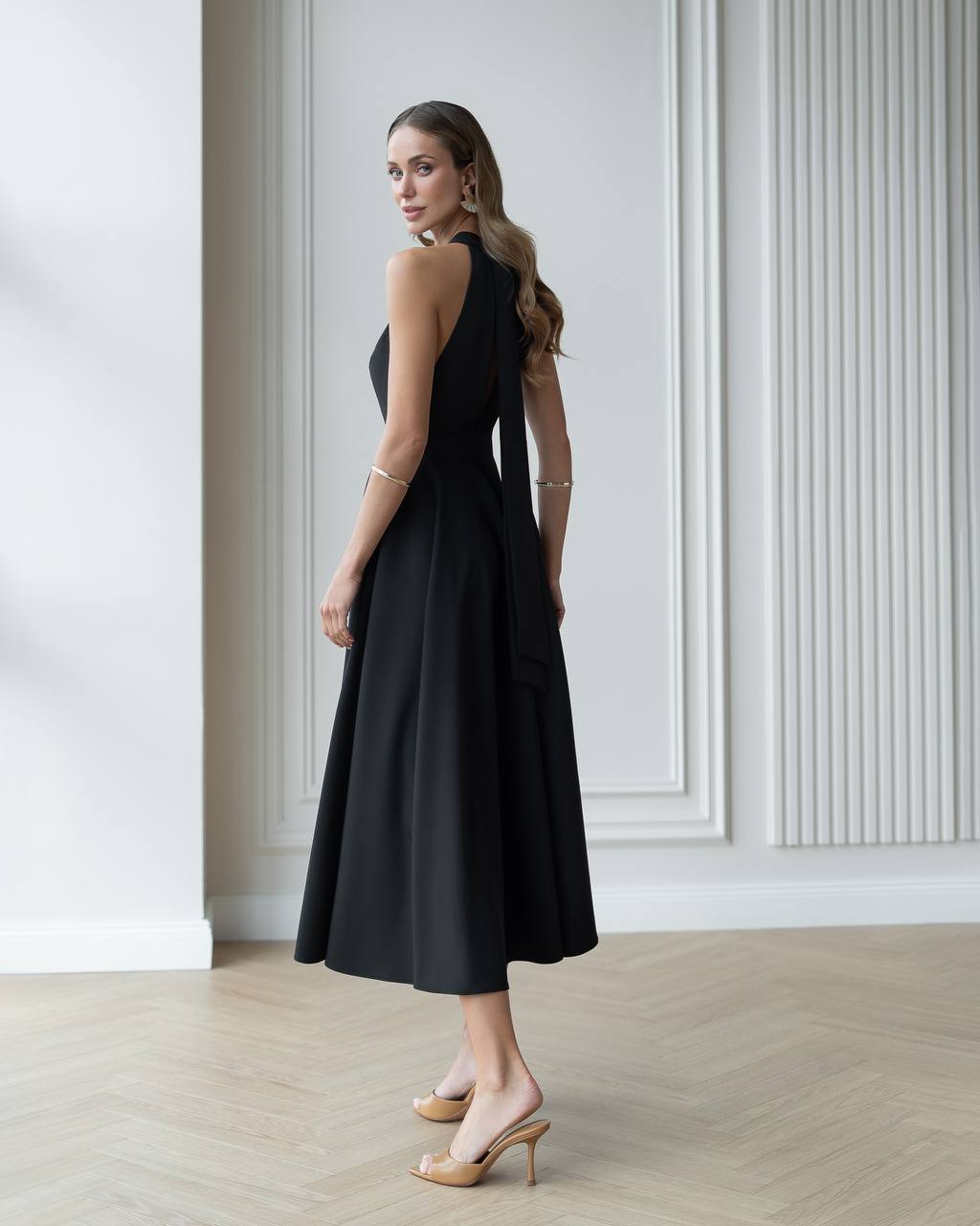 Woman wearing a black dress standing in a room with white walls and wooden flooring.