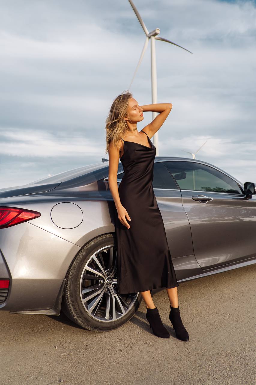 Woman in a black dress standing next to a silver car with a wind turbine in the background