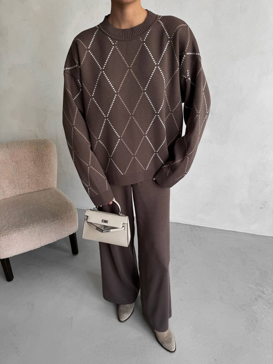 Person wearing a brown diamond-patterned sweater and matching pants, holding a beige handbag against a neutral background.