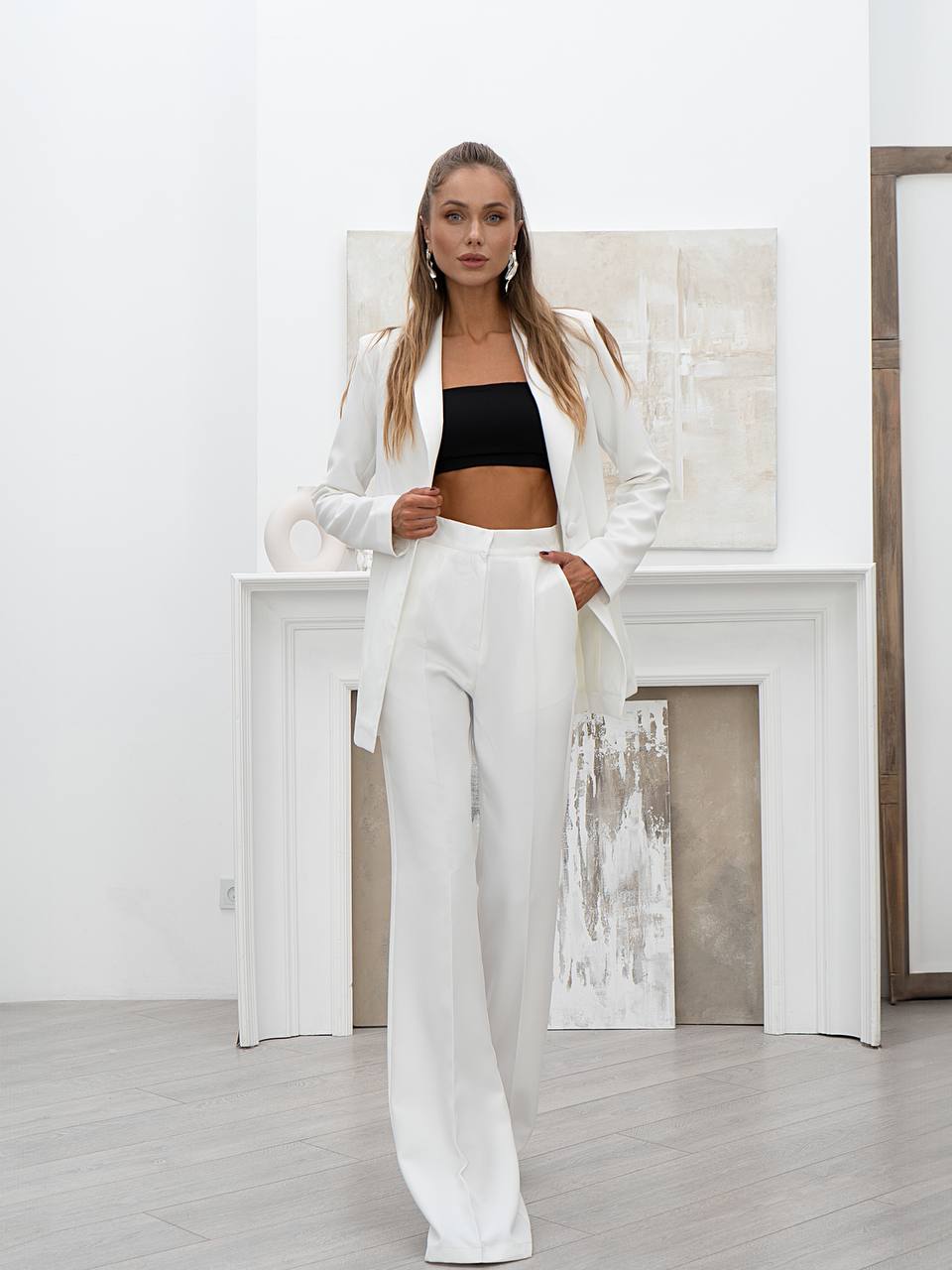 Woman in a white suit standing in a room with a fireplace and abstract painting.