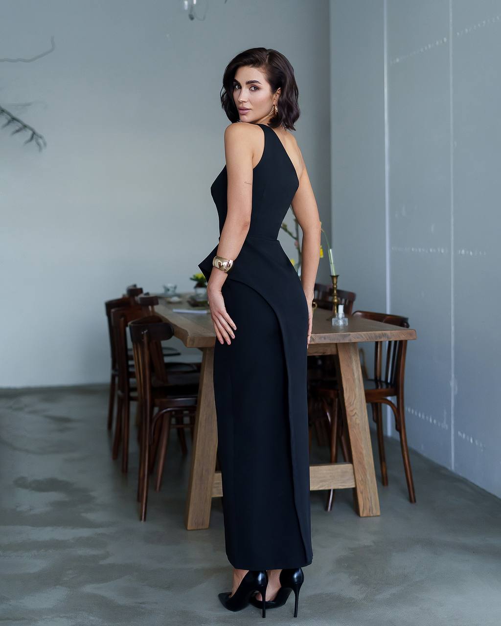 Woman in a black dress standing in a modern interior setting with a dining table.