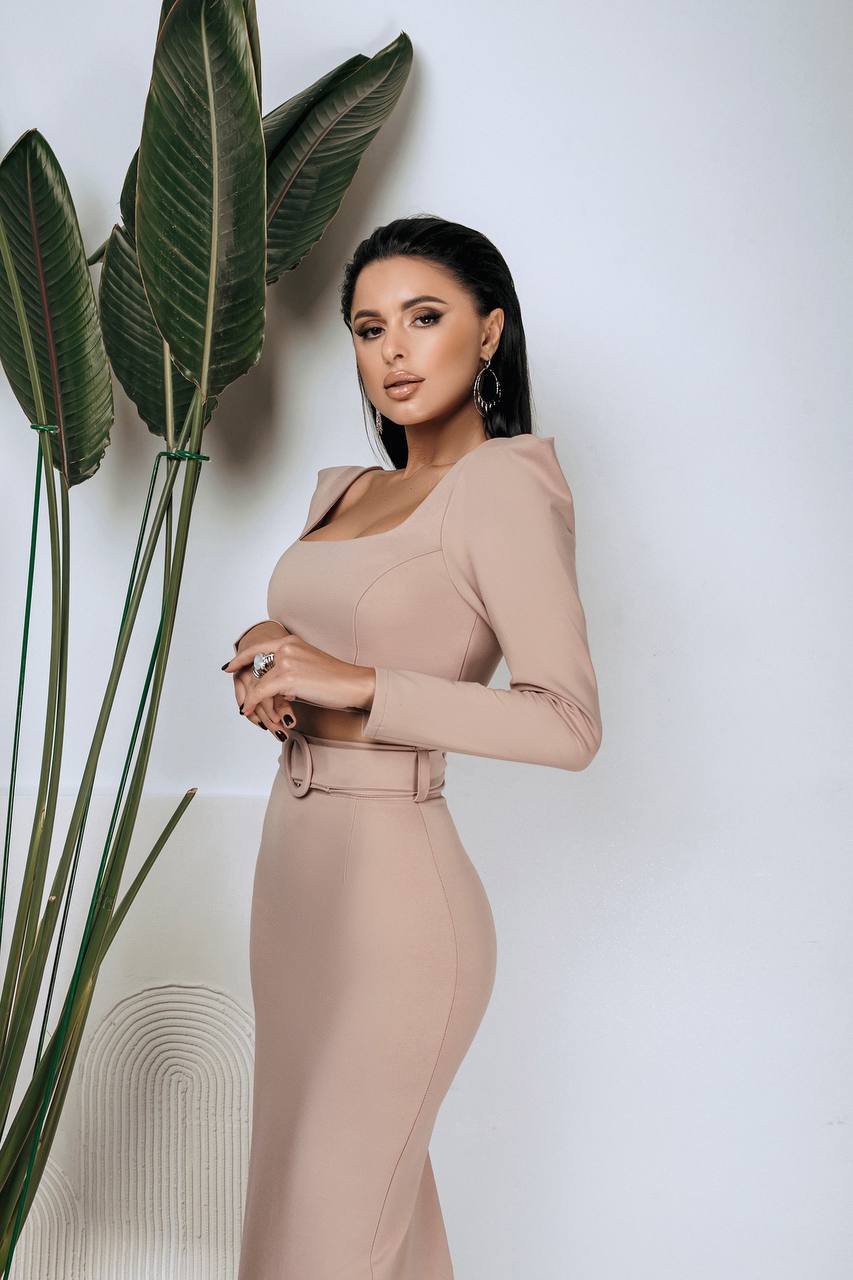 Woman in a beige dress standing next to a plant against a white wall