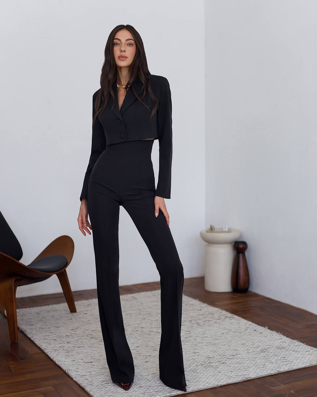 Woman wearing a black suit standing in a minimalistic room with a white wall and wooden floor.