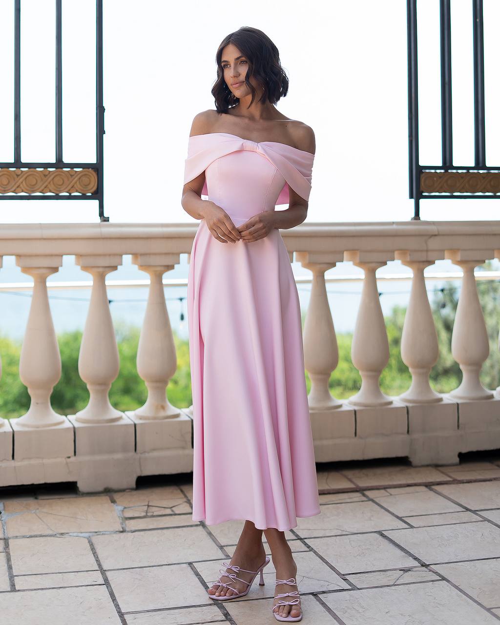 Woman in a pink off-shoulder dress standing on a balcony with a scenic background
