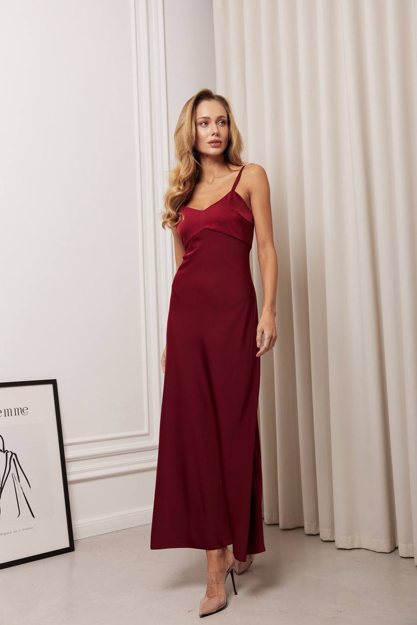 Woman wearing a burgundy dress standing in a room with a white wall and beige curtain.