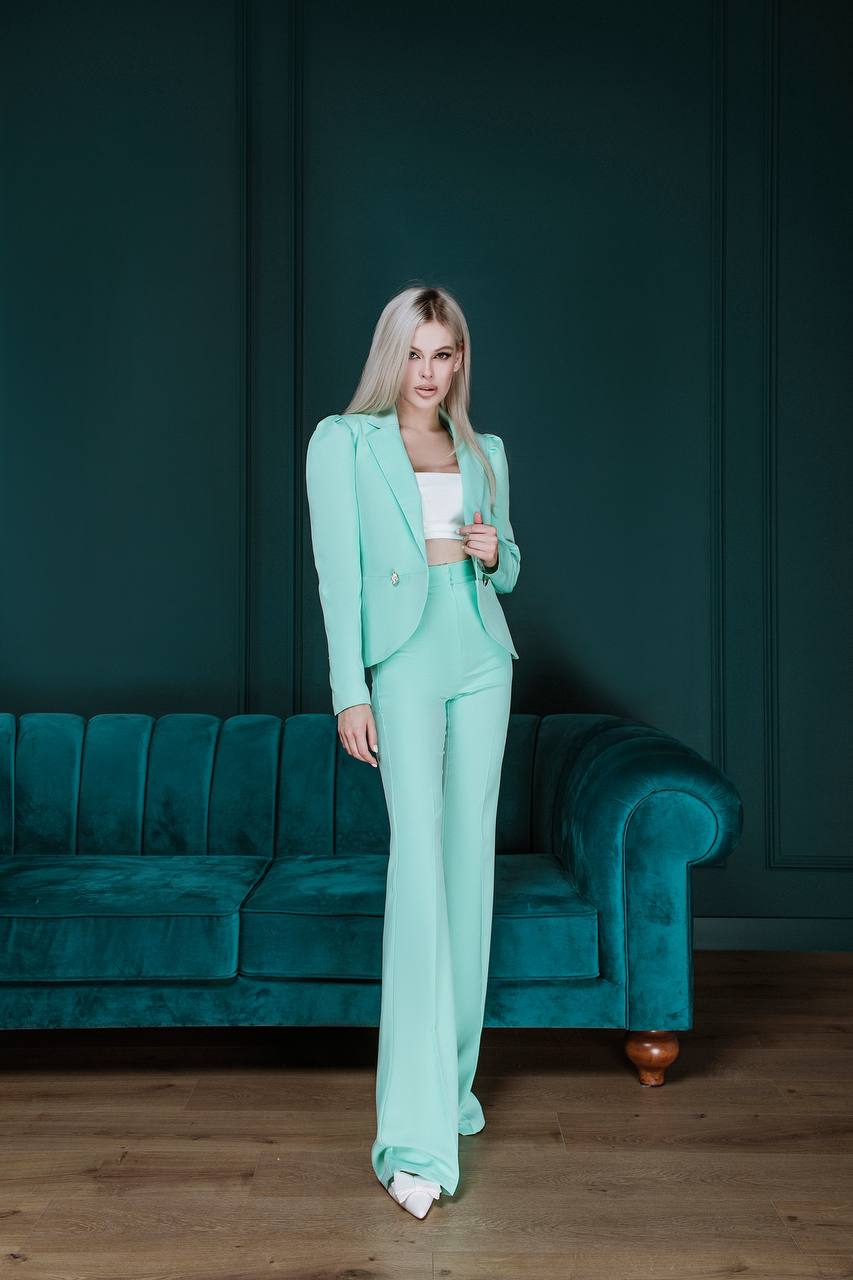 Woman in a mint green suit standing in a room with a teal couch and dark walls.