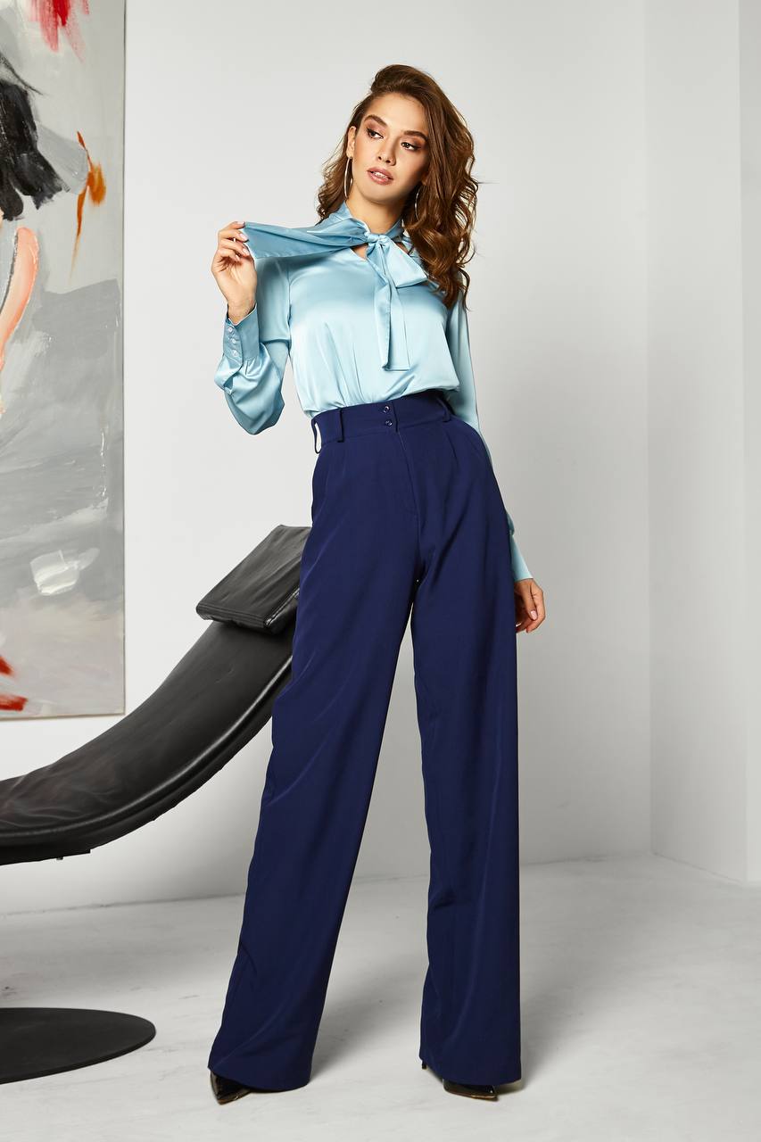 Woman wearing a light blue blouse and dark blue pants in a minimalistic setting.