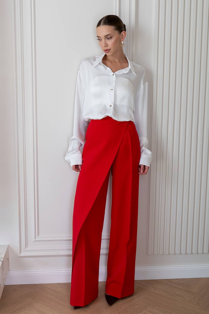 Woman wearing a white blouse and red pants against a white wall.