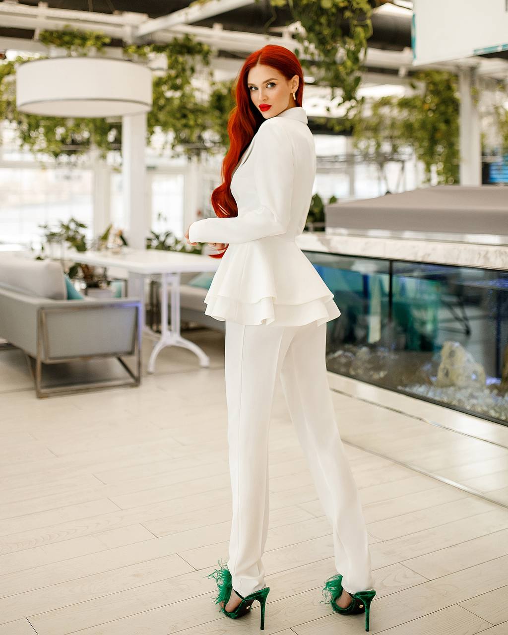 Woman in a white suit with green shoes standing in an indoor setting with plants.