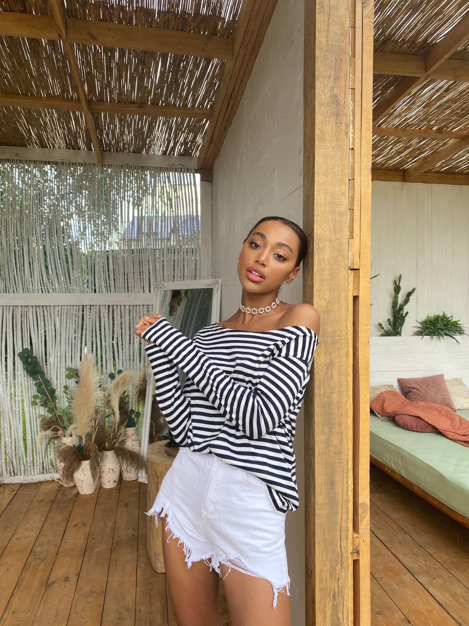 Woman in a striped top and white shorts standing in a wooden structure with plants and a bed in the background.