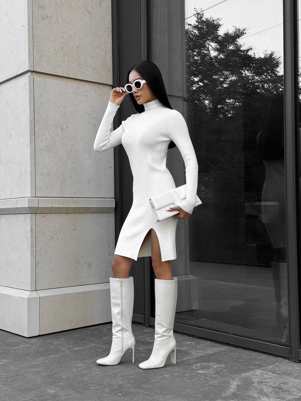 Woman in a white dress and knee-high boots standing outside a building.