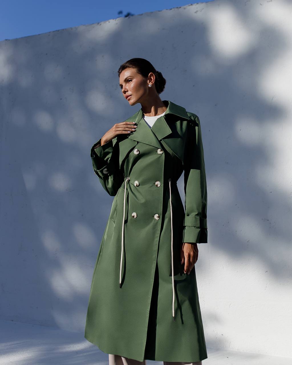 Woman wearing a green trench coat against a white wall with blue sky.