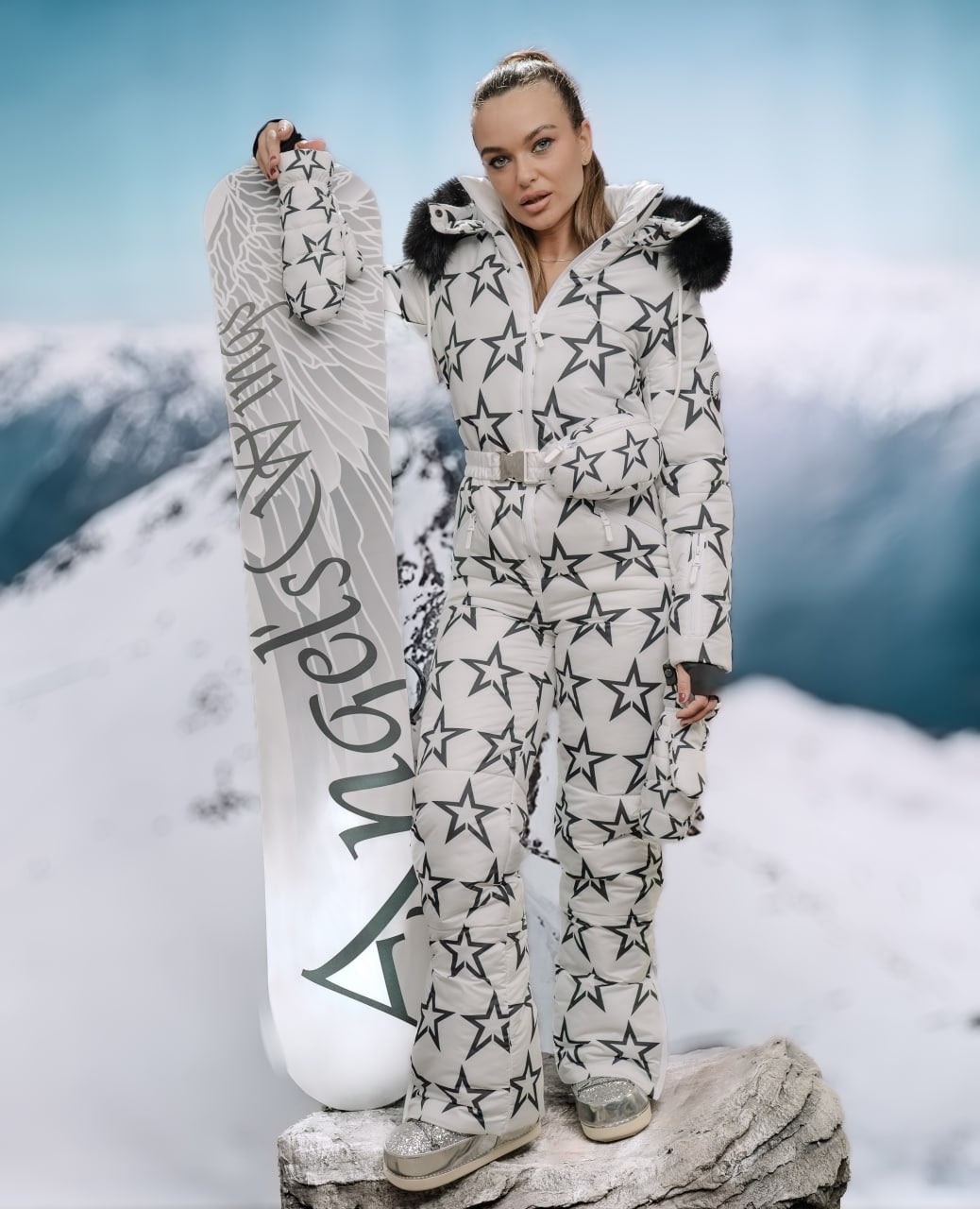 Person in star-patterned snowsuit holding a snowboard with mountains in the background