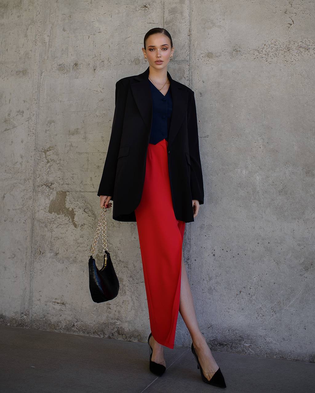 Woman in a black blazer, navy top, and red skirt standing against a concrete wall.