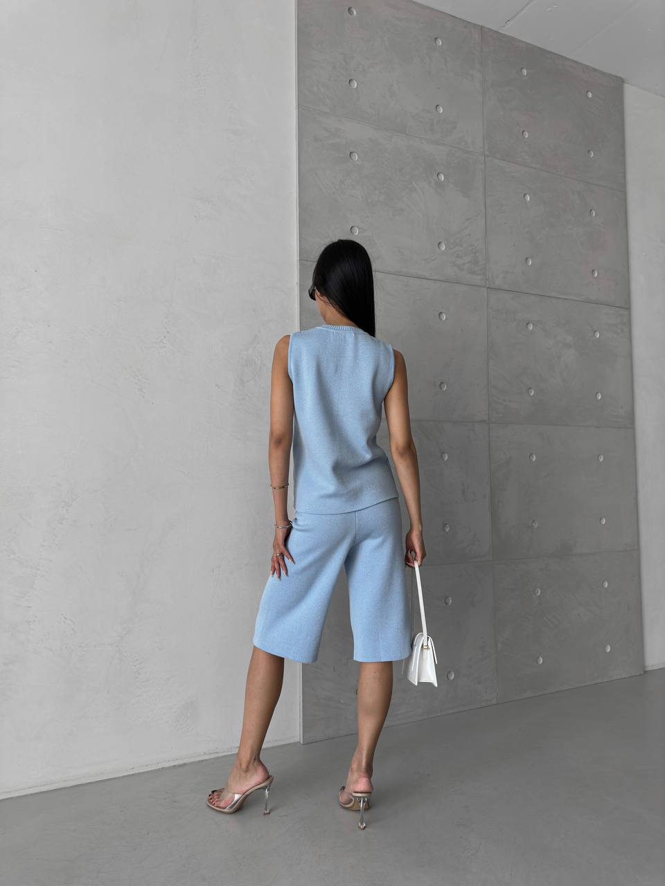 Woman in a light blue sleeveless top and shorts standing against a gray concrete wall.
