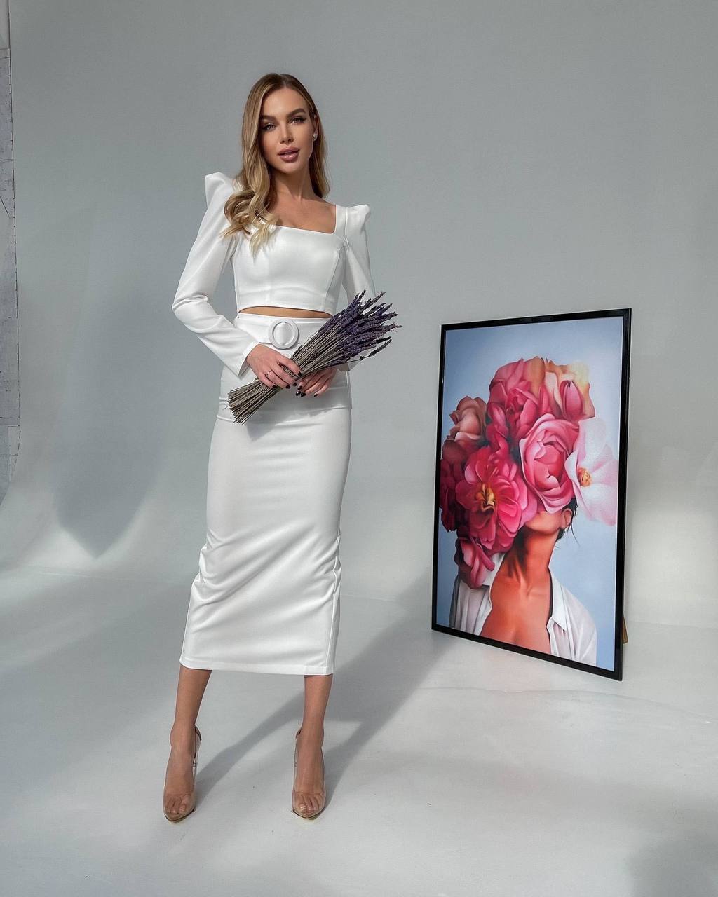 Woman in a white outfit holding flowers next to a framed artwork on a gray background