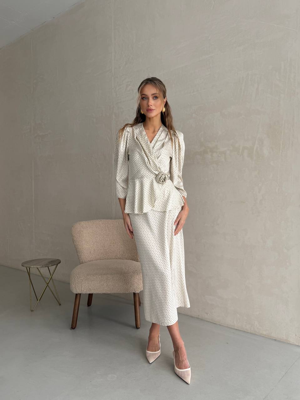 Woman in a white patterned outfit standing in a minimalistic room with a chair and small table.