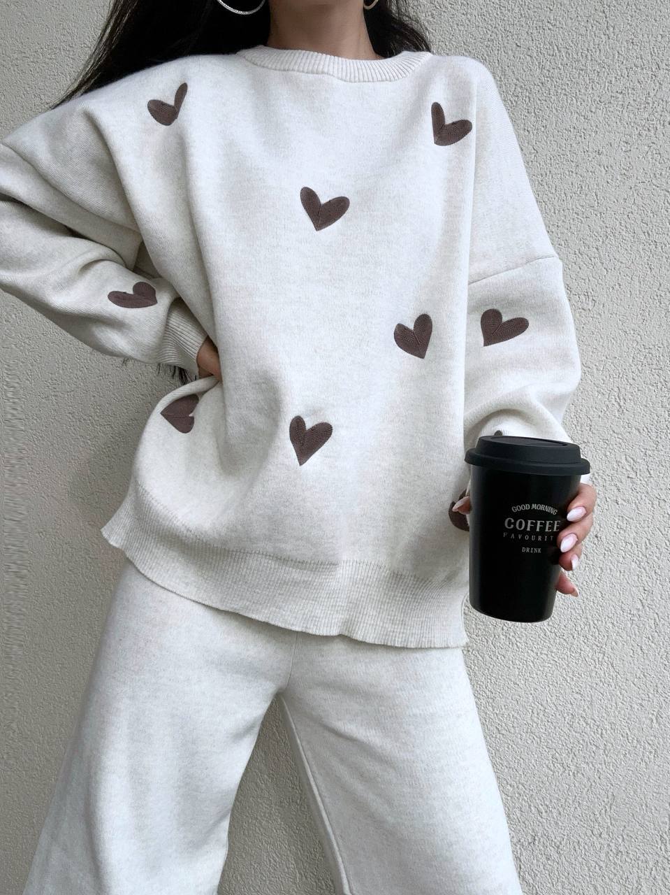 Person wearing a white sweatshirt with brown heart patterns, holding a black coffee cup against a plain background.