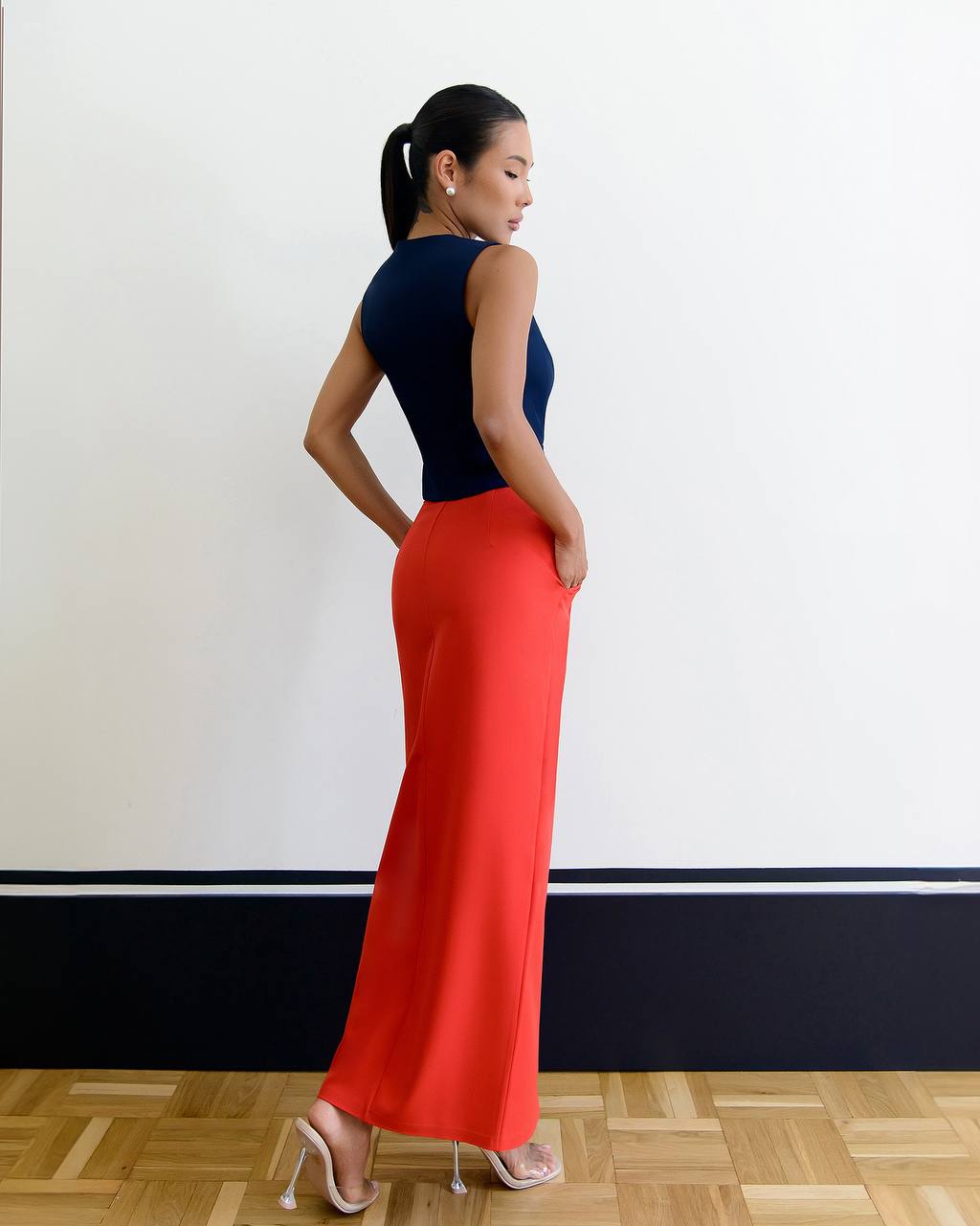 Woman wearing a black top and red pants standing against a white wall.