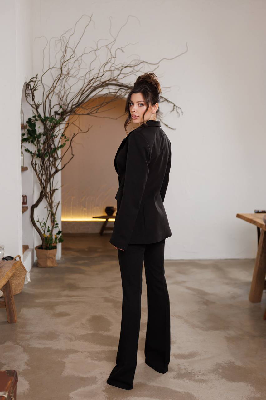 Woman in a black outfit standing in a room with a plant and wooden furniture.