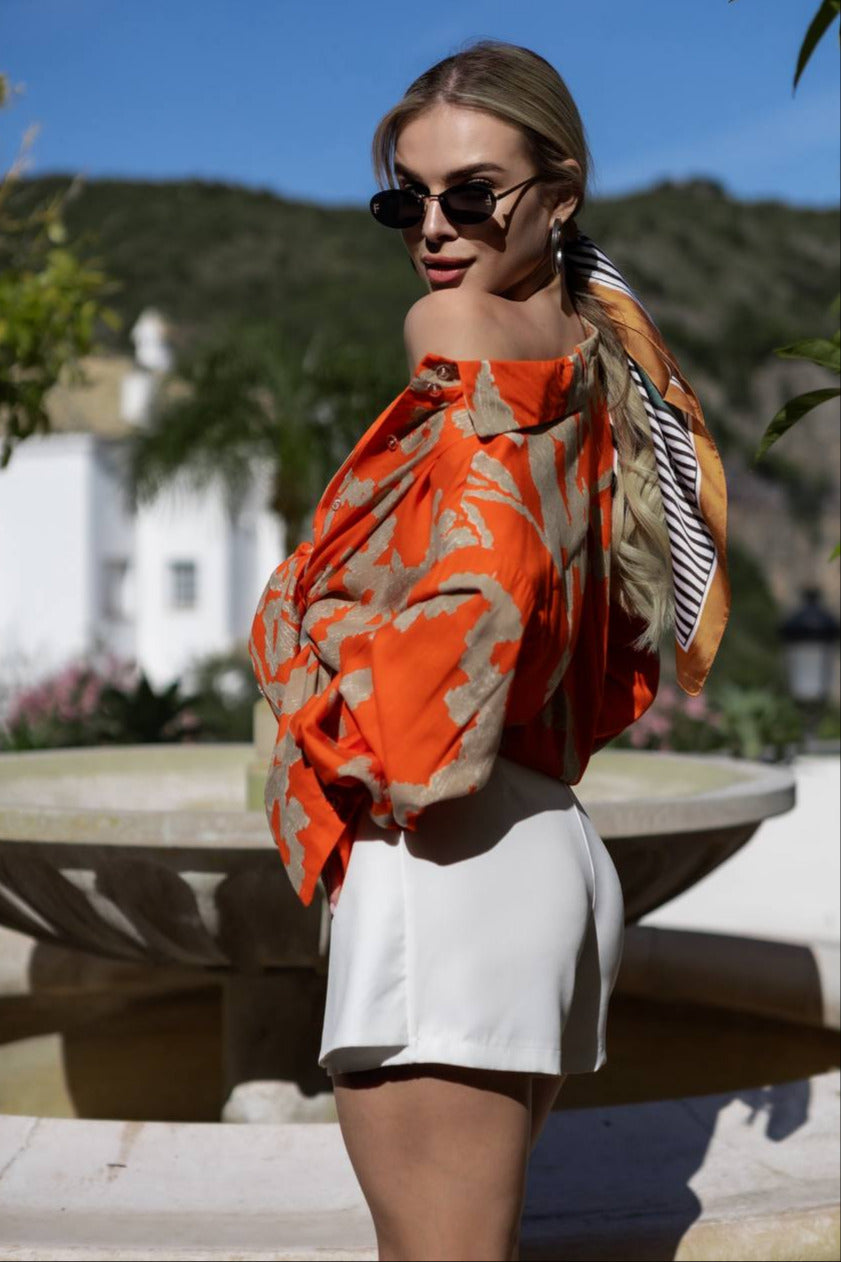 Woman wearing an orange floral off-shoulder top and white shorts outdoors.