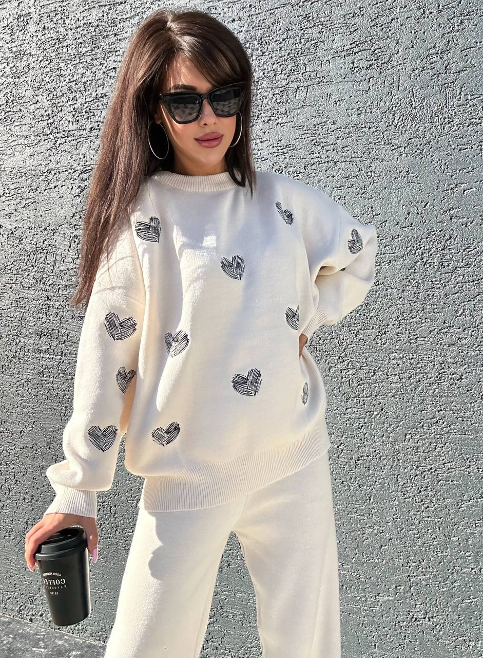 Woman wearing a white sweater and pants with heart patterns, holding a black coffee cup against a gray wall.