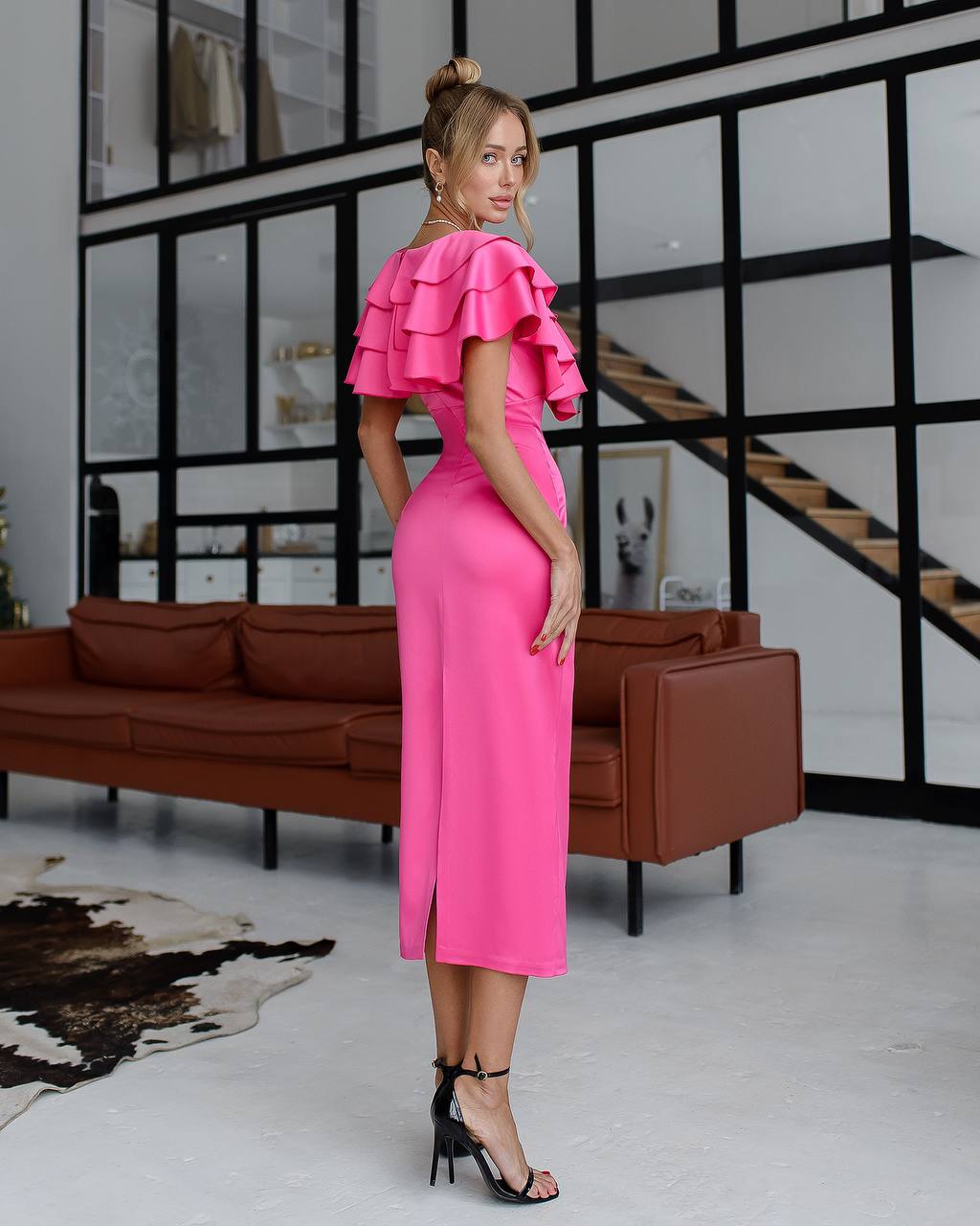 Woman in a bright pink dress standing in a modern interior setting with a brown sofa and glass wall.