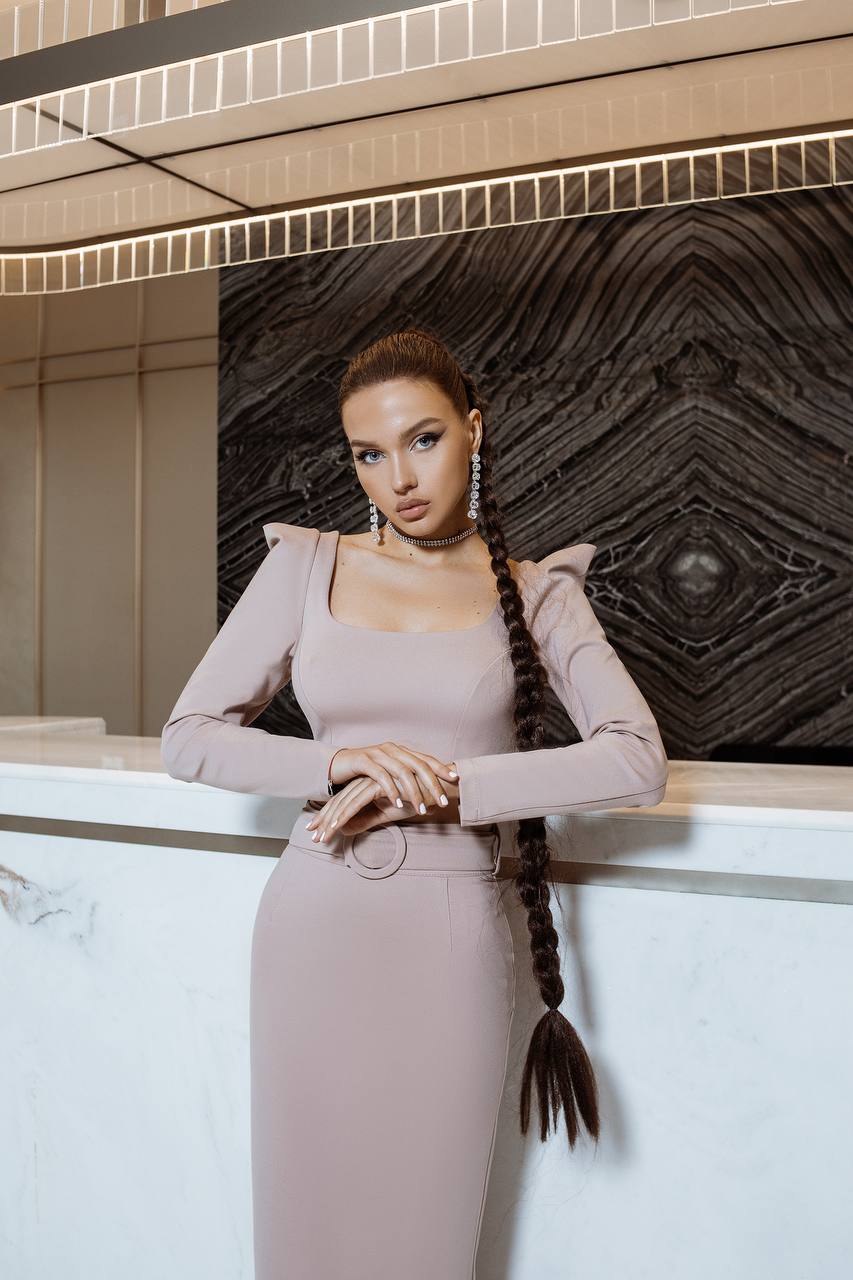 Woman in a light pink dress with braided hair standing against a decorative wall.