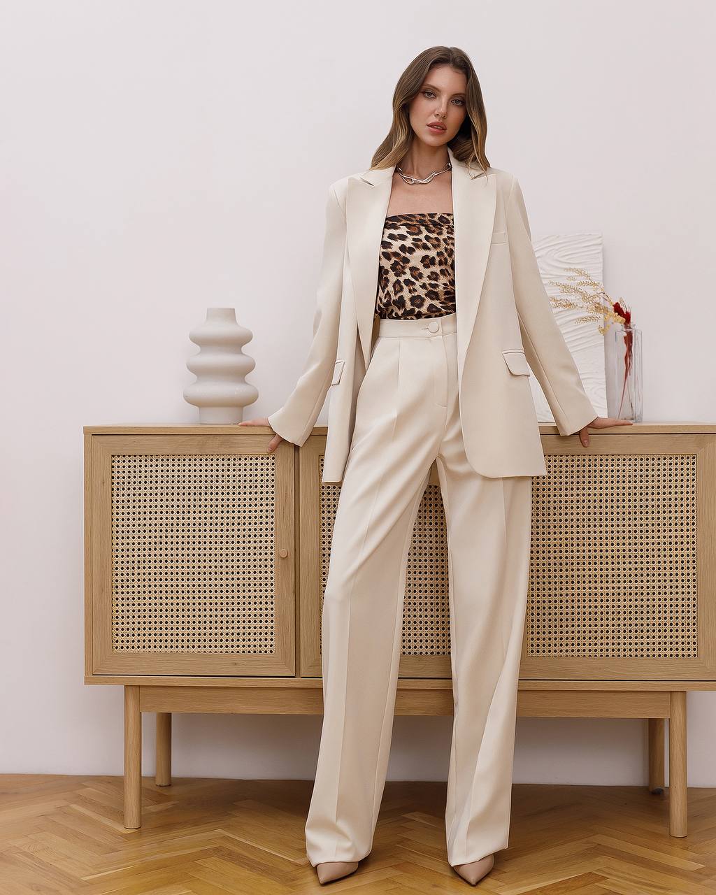 Woman in a beige suit with a leopard print top standing in front of a wooden cabinet.