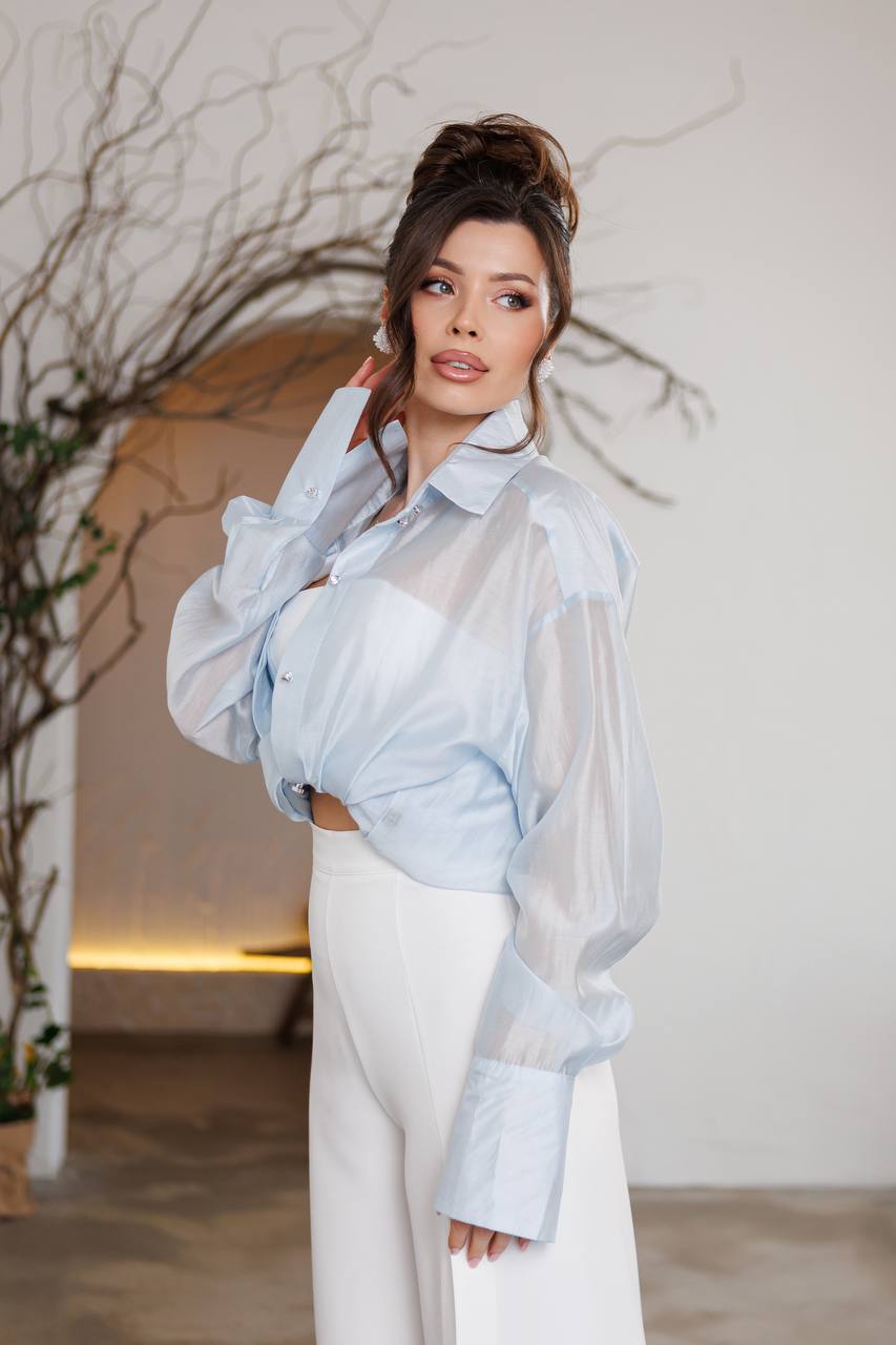 Woman wearing a light blue sheer blouse over a white outfit with a neutral background