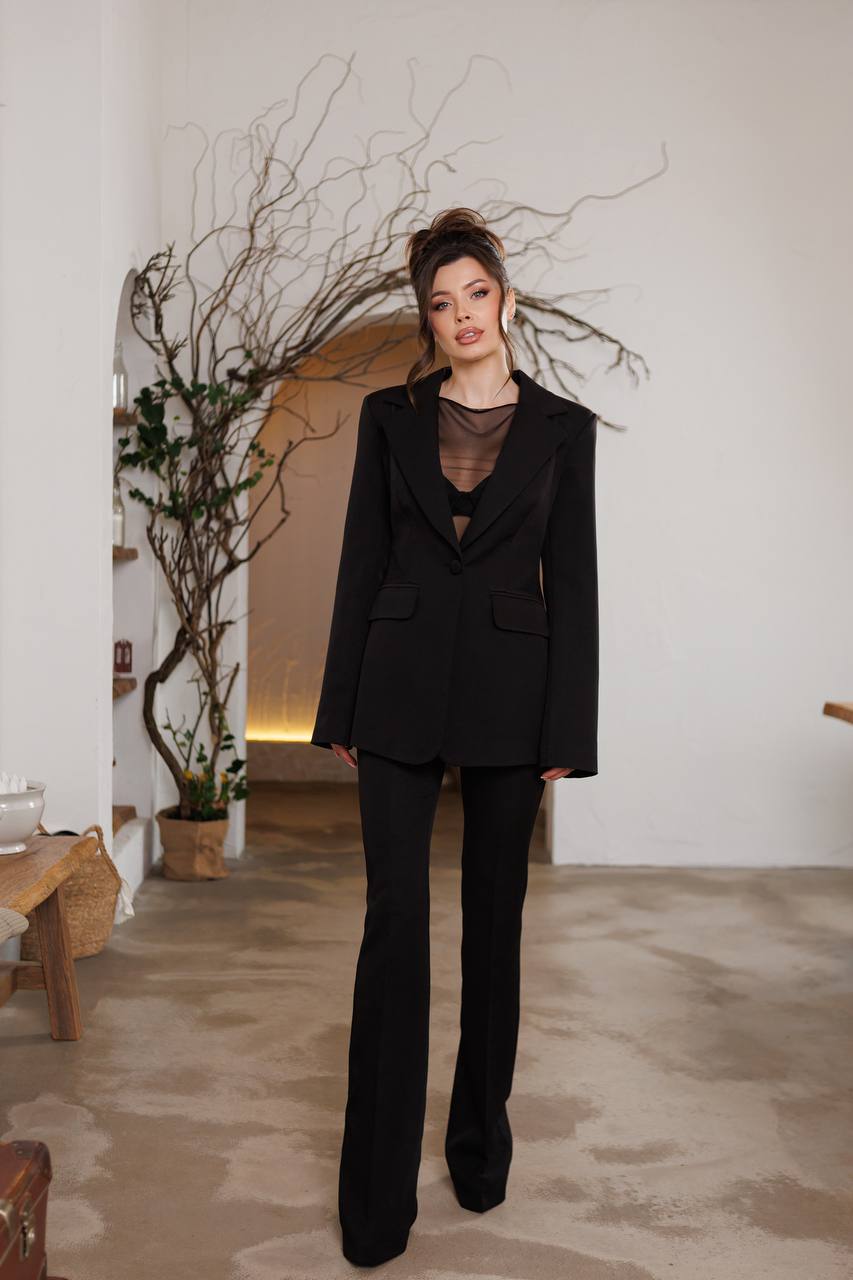 Woman in a black suit standing in an indoor setting with minimal decor.