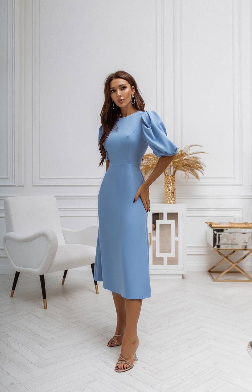 Woman in a light blue dress standing in a stylish room with white walls and decor.
