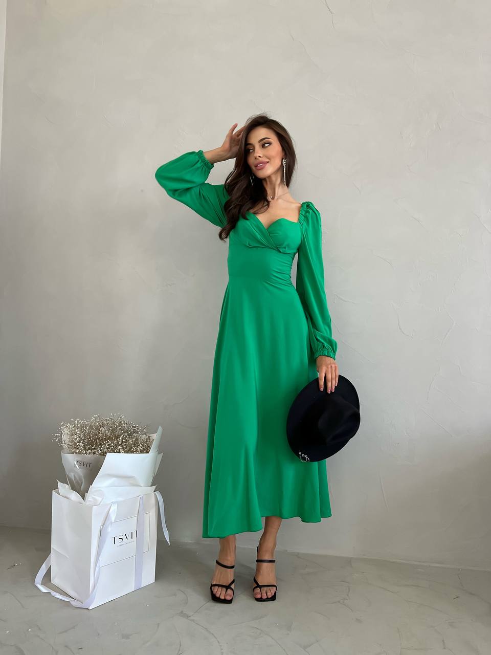 Woman in a green dress holding a black hat, standing next to a white bag with 'Dior' branding.