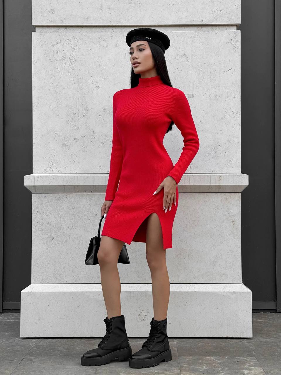 Person wearing a red dress with a black beret and boots standing against a gray wall.
