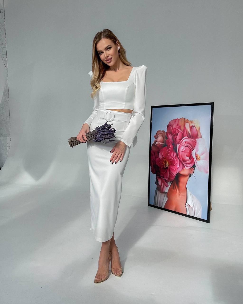 Woman in a white outfit holding flowers next to a framed artwork on a gray background