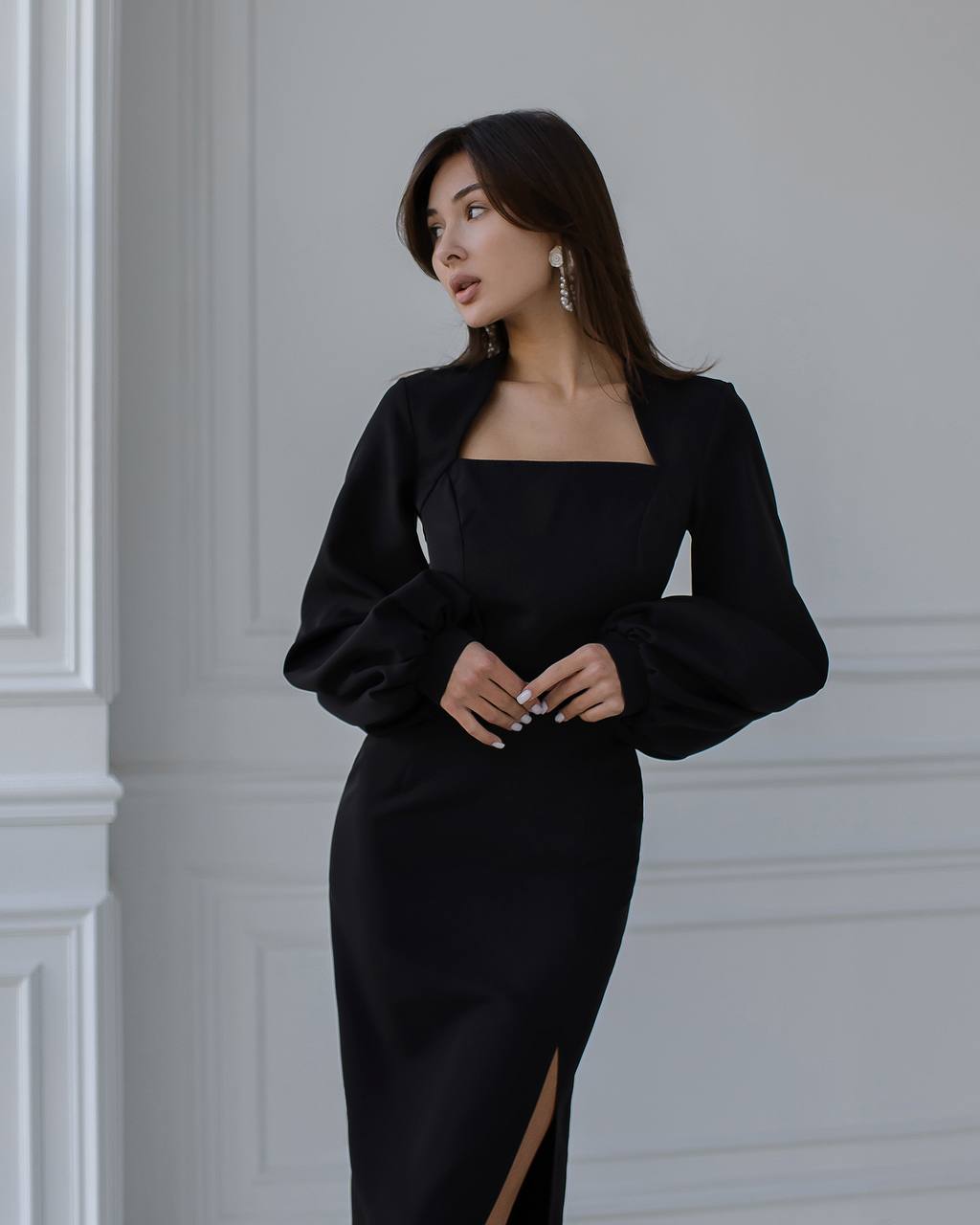 Woman wearing a black dress with a slit, standing against a gray wall.