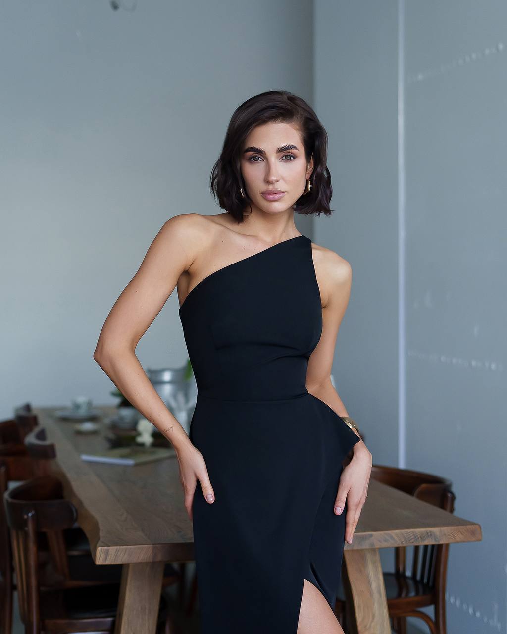 Woman in a black one-shoulder dress standing in a room with wooden furniture.