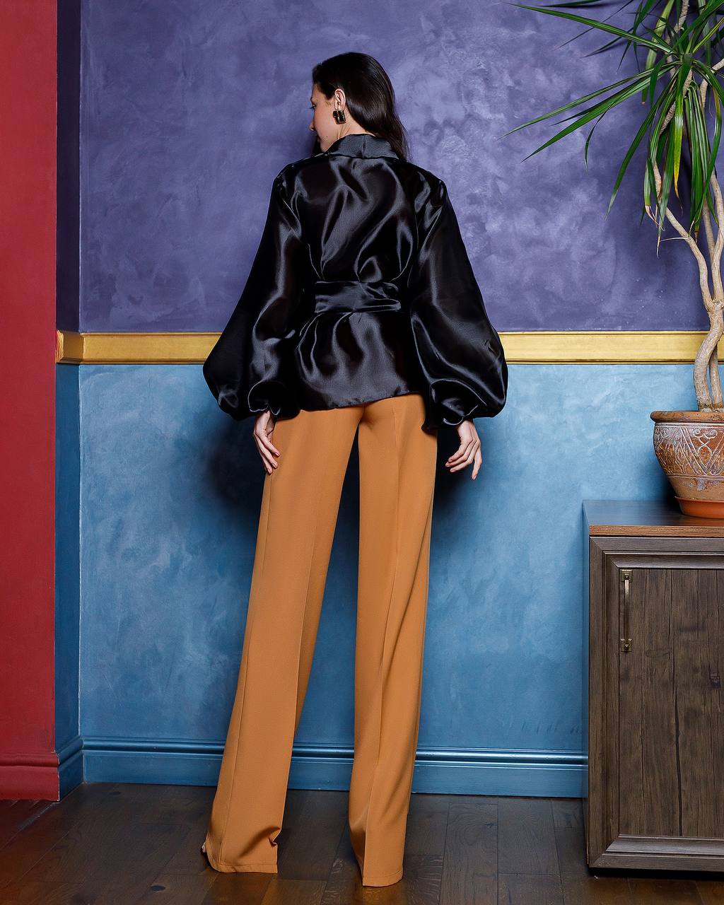Person wearing a black satin jacket and tan pants against a colorful wall with a plant and cabinet.