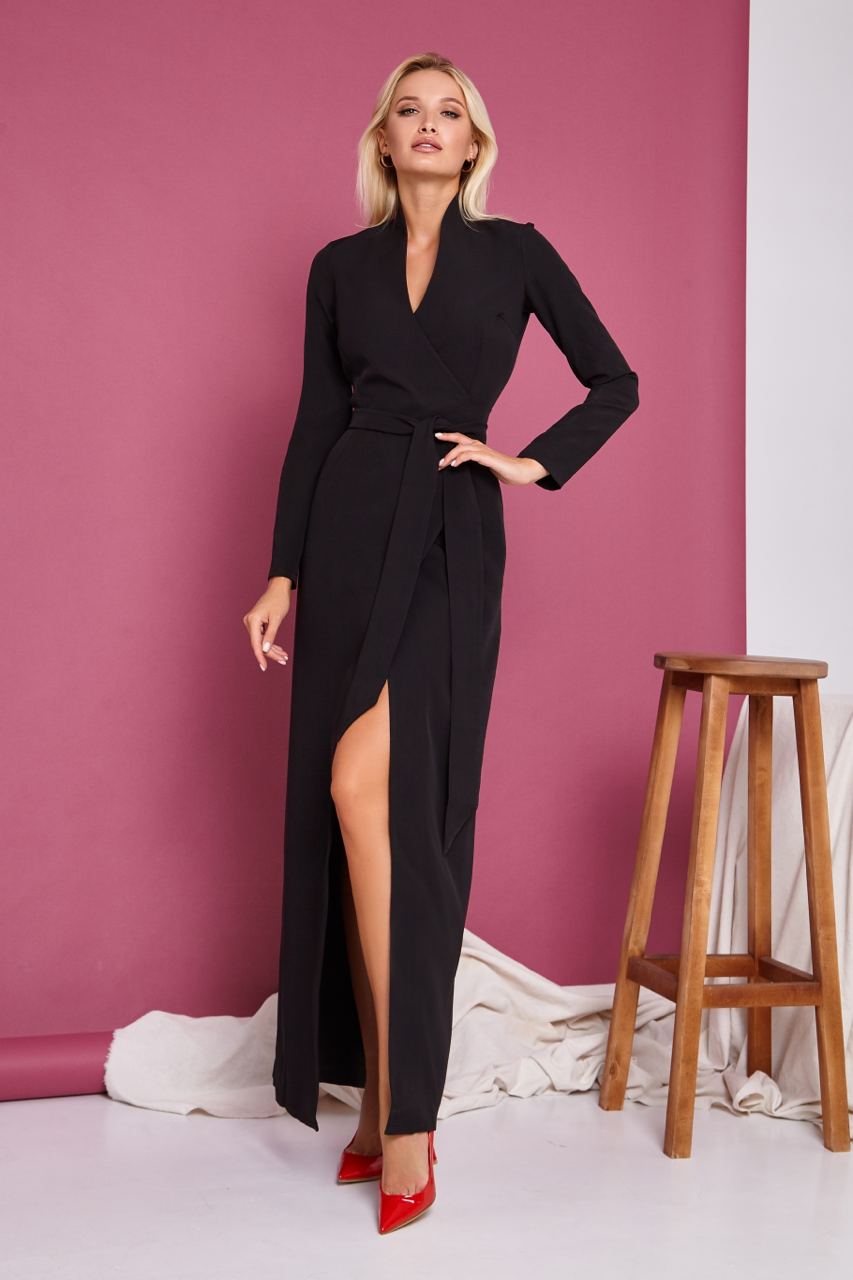 Woman wearing a black dress with a high slit, standing against a pink wall with a wooden stool.